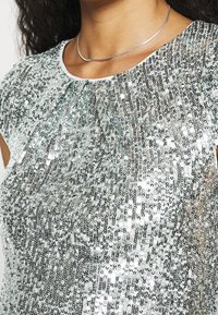 Haut en sequins argentés avec un col froncé. Texture réfléchissante et scintillante, manches courtes et doublure lisse en dessous.