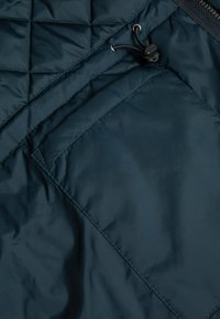 Námorní modrá prošívaná látka s hladkým povrchem, obsahující kapsu na zip a stahovací šňůrku nahoře.