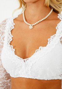 Weißes Spitzen-Bralette mit gewellten Kanten, das florale Muster zeigt. Eine Perlenkette mit einem herzförmigen Anhänger ist sichtbar.