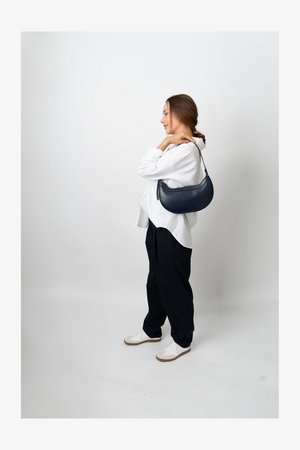 Námornícka kožená taška na rameno s zaobleným tvarom, hladkým povrchom a jedným popruhom, nosená proti bielej košeli a pruhovaným nohaviciam.