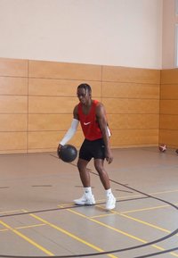 Jugador de baloncesto con camiseta sin mangas roja y pantalones cortos negros, sosteniendo un balón de baloncesto negro. El fondo presenta paneles de madera y líneas de la cancha.