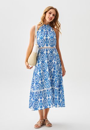 Biała sukienka midi z niebieskimi kwiatowymi wzorami, z dekoltem halter i falbanami. Noszona z beżowymi sandałami na paski i plecioną torebką.