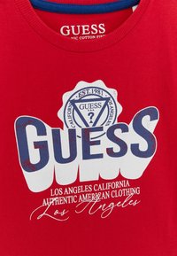 Rød bomulds t-shirt med en stor hvid og blå "GUESS" grafik, med et logo og mindre tekst, der siger "Los Angeles California."