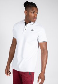 Witte poloshirt met zwarte accenten, knoopsluiting met drie knopen en geborduurd logo. Korte mouwen met contrasterende manchetten. Draagt het met rode textuurbroek.