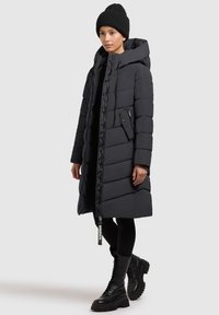 Czarna ocieplana kurtka z kapturem, zamkiem z przodu, dużą boczną kieszenią i dzianinową czapką. Modelka ma na sobie czarne leginsy i sznurowane botki.