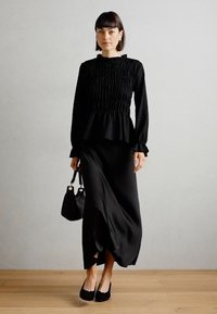Black peplum top with smocked texture and long sleeves, paired with a flowing black maxi skirt. Accessorized with a small handbag and flats.