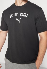 Černé bavlněné tričko s bílým nápisem "FC ST. PAULI" a bílým skákajícím logem pumy, krátké rukávy a kulatý výstřih.