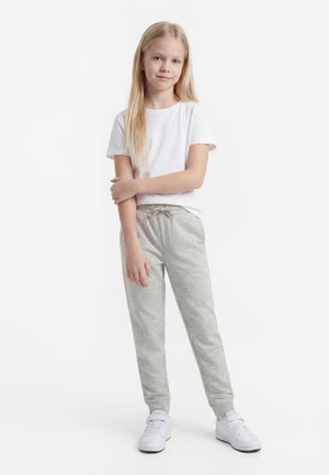 Ξανθιά κοπέλα στέκεται με σταυρωμένα χέρια, φοράει λευκό μπλουζάκι, ανοιχτό γκρι φόρμα και λευκά αθλητικά παπούτσια καθώς στέκεται σε λευκό φόντο.