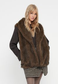 Blonde vrouw die een donkerbruin nepbont vest over een zwarte lange mouwen shirt en een geruite mini rok draagt, poseert met één hand in haar zak.
