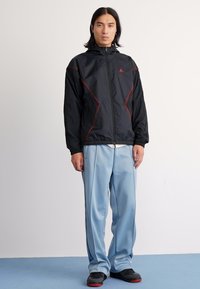 Ung mand står iført en sort lynlåsjakke med røde detaljer, løse lyseblå bukser og sorte sneakers på blå gulv.