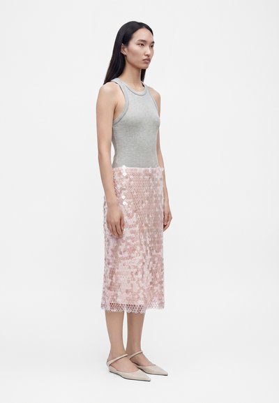 Femme debout, tournée vers la droite, portant un haut gris sans manches et une jupe midi rose à sequins avec des ballerines beiges à bouts pointus sur fond blanc uni.