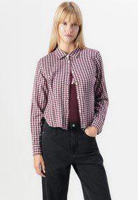Red and white checkered button-up shirt with a collar, paired with a maroon top and high-waisted black jeans. Soft fabric, structured fit.