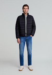 Sort pufferjakke med vandret quiltet tekstur, frontlampe og et lille logo. Sammen med lyseblå jeans og hvide sneakers.