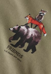 Camiseta con un diseño impreso de un oso, un zorro y un mapache sobre un tejido texturizado de color verde oliva, con el texto "AMIGOS en todas formas y tamaños".