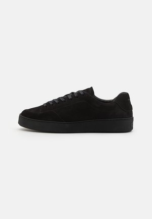 Black suede sneakers with a rounded toe, flat laces, and a textured black rubber sole, featuring a minimalist design and no prominent logos.