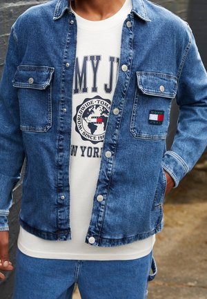 Person iført en blå denimjakke over en hvid grafisk t-shirt med en globus og tekst, kombineret med blå denimbukser.