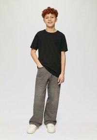 Rothaariger Junge steht mit einer Hand in der Tasche, trägt ein schwarzes T-Shirt, graue weit geschnittene Jeans und weiße Sneaker, und lächelt leicht.
