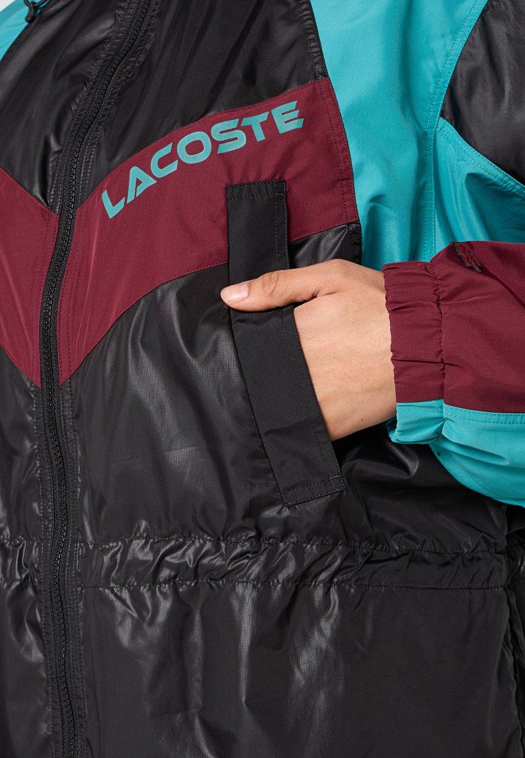 Czarna i turkusowa kurtka przeciwdeszczowa z bordowymi akcentami, zapinana na zamek z przodu i z elastycznymi mankietami; zawiera boczną kieszeń z widocznym detalem szycia.