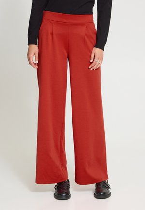 Femme portant un pantalon large taille haute couleur rouille, un haut noir à manches longues et des chaussures en cuir noires, debout sur un sol clair.