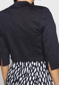 Marineblauwe cropped jas met korte mouwen en gespleten manchetten, gemaakt van een gladde stof, gecombineerd met een zwart-witte gestreepte rok.