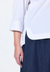 Blusa di cotone bianca con orlo arrotondato e maniche a tre quarti con polsini. Abbinata a una gonna in denim blu scuro, che mostra una texture liscia.