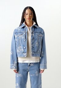 Veste en denim bleu clair avec des détails déchirés, col arrondi et fermeture à boutons, associée à un sweat à capuche blanc zippé et un jean assorti.
