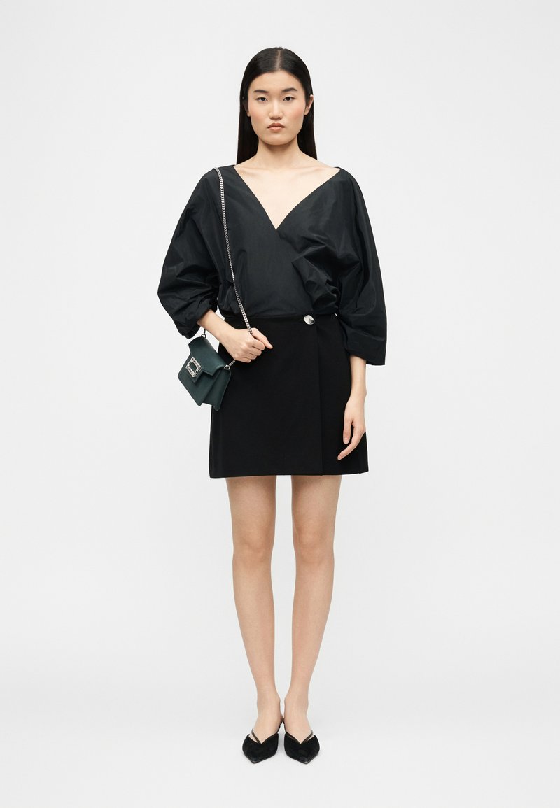 Γυναίκα με μακριά σκούρα μαλλιά που φοράει μαύρο μπλουζάκι με V λαιμόκοψη και φουσκωτά μανίκια, μαύρη μίνι φούστα, μαύρα επίπεδα παπούτσια και μια μικρή σκοτεινή πράσινη τσάντα.