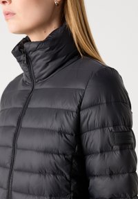 Veste rembourrée noire avec un col haut, des coutures horizontales et une finition lisse. Présente un patch logo sur la manche et une fermeture éclair.