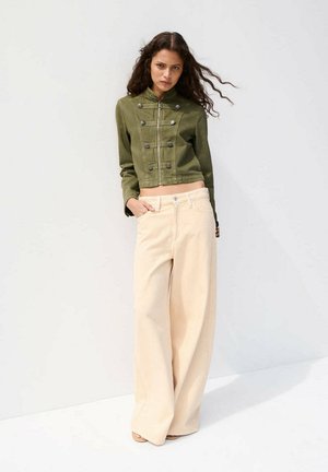 Mujer con cabello largo y rizado que lleva una chaqueta corta verde oliva y pantalones beige de talle alto y pierna ancha, de pie contra una pared blanca.