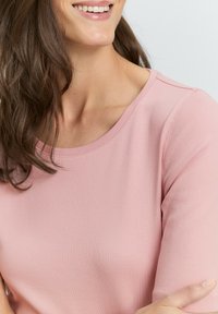 Světle růžový top s krátkým rukávem a kulatým výstřihem, vyrobený z měkké, texturované látky. Obsahuje viditelné švy podél švů.