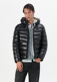 Schwarze Steppjacke mit glänzender, gesteppten Textur, Kapuze und Frontreißverschluss. Kombiniert mit einem hellblauen Hemd und dunklen Jeans. Mit sichtbarem Logo.