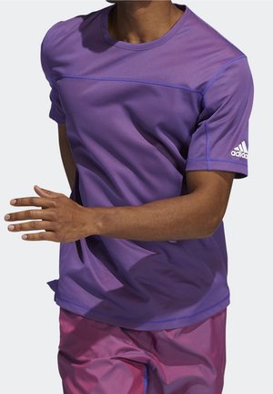 Homme courant portant un t-shirt à manches courtes violet Adidas et un short de sport rose, visible du cou jusqu'au milieu de la cuisse sur un fond uni.