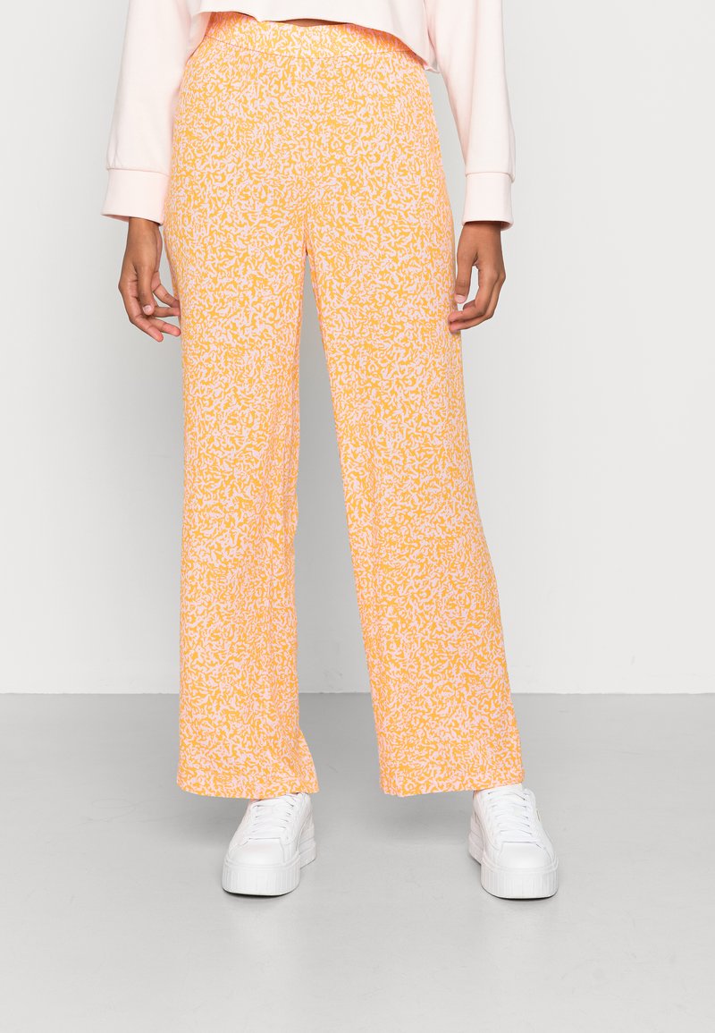 Calças largas laranja apresentam um padrão texturizado com detalhes brancos. Feitas de um tecido leve, combinadas com tênis brancos.