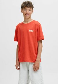 Camiseta de algodón roja con cuello redondo y mangas cortas, que presenta un logo blanco de "SAINT-TROPEZ" en el pecho. Se lleva con pantalones cortos de denim claros.