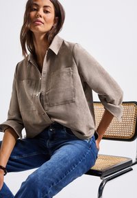 Frau mit schulterlangem Haar, die ein beiges Cordhemd und eine blaue Jeans trägt, sitzt auf einem Stuhl mit einem Gestell aus Rattan und Metall vor weißem Hintergrund.