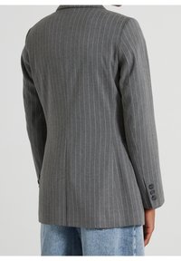 Blazer gris à fines rayures avec poignets à trois boutons, porté avec un jean bleu clair, vu de dos sur un fond clair uni.