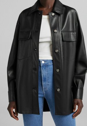 Personne portant une veste-chemise en cuir noir boutonnée sur un haut blanc côtelé et un jean bleu taille haute sur fond gris.
