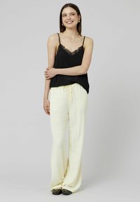 Camisole noire avec bordure en dentelle, à fines bretelles, associée à un pantalon léger jaune pâle à cordon de serrage. Modèle portant des chaussures noires à enfiler.