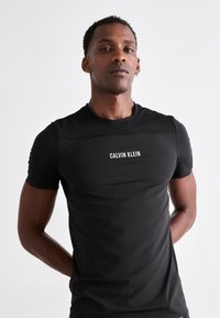 Man wearing a fitted black Calvin Klein t-shirt, standing with hands behind back against a plain white background.