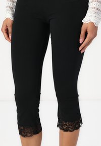 Leggings capri noirs avec une texture lisse, dotés d'une finition en dentelle festonnée à l'ourlet. Longueur mi-mollet et coupe extensible.