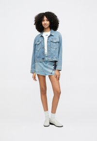 Τζιν μπουφάν με δύο τσέπες στο στήθος, συνδυασμένο με μια ανοιχτό μπλε φούστα με γραφικό σχέδιο και ξεφτισμένο τελείωμα. Το μοντέλο φοράει λευκές κάλτσες και παπούτσια.