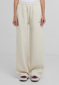 Pantaloni della tuta a gamba larga, di colore crema, con vita elasticizzata e logo ricamato rosa sulla coscia. Il materiale sembra morbido e strutturato.