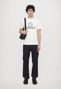 Hvid bomulds T-shirt med "VERSACE" tryk og logo, parret med mørke denim jeans, sort skuldertaske og sorte sko.
