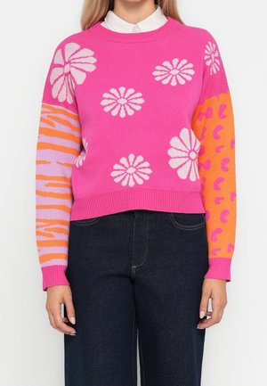 Pull rose vif avec de grands motifs de marguerites blanches, une manche orange avec des taches abstraites roses, et une manche violette avec des rayures orange.
