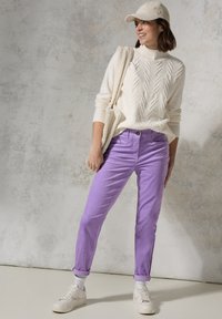 Wit gebreid vest met een textuurpatroon, lichtpaarse broek opgerold bij de manchetten, witte sneakers en een beige pet, staande tegen een gedempte achtergrond.