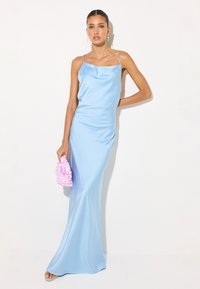 Femme portant une robe nuisette en satin bleu clair tenant une pochette rose texturée, debout devant un fond blanc uni.