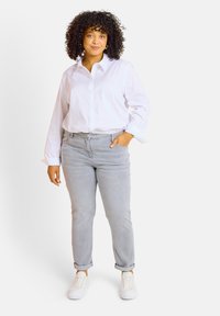 Camisa blanca de botones, jeans gris claro y zapatillas blancas. El modelo se encuentra de pie con una mano en el bolsillo, exhibiendo un ajuste casual y relajado. Cabello rizado.