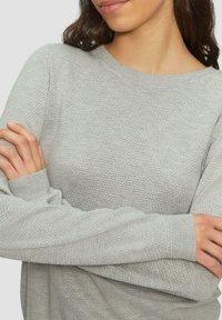 Hellgrauer Pullover mit strukturiertem Wabenmuster, Rundhalsausschnitt und gerippten Bündchen. Nahaufnahme, die die Details des Stoffs und die Passform betont.