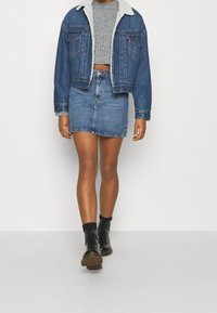 Mujer vestida con un suéter gris corto, chaqueta de mezclilla azul con cuello de forro blanco, falda de mezclilla a juego, calcetines negros y botas negras con cordones.