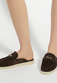 Brune suède loafers met een open achterkant, voorzien van een gouden accent en een crèmekleurige rubberen zool. Smooth textuur, klassieke design.